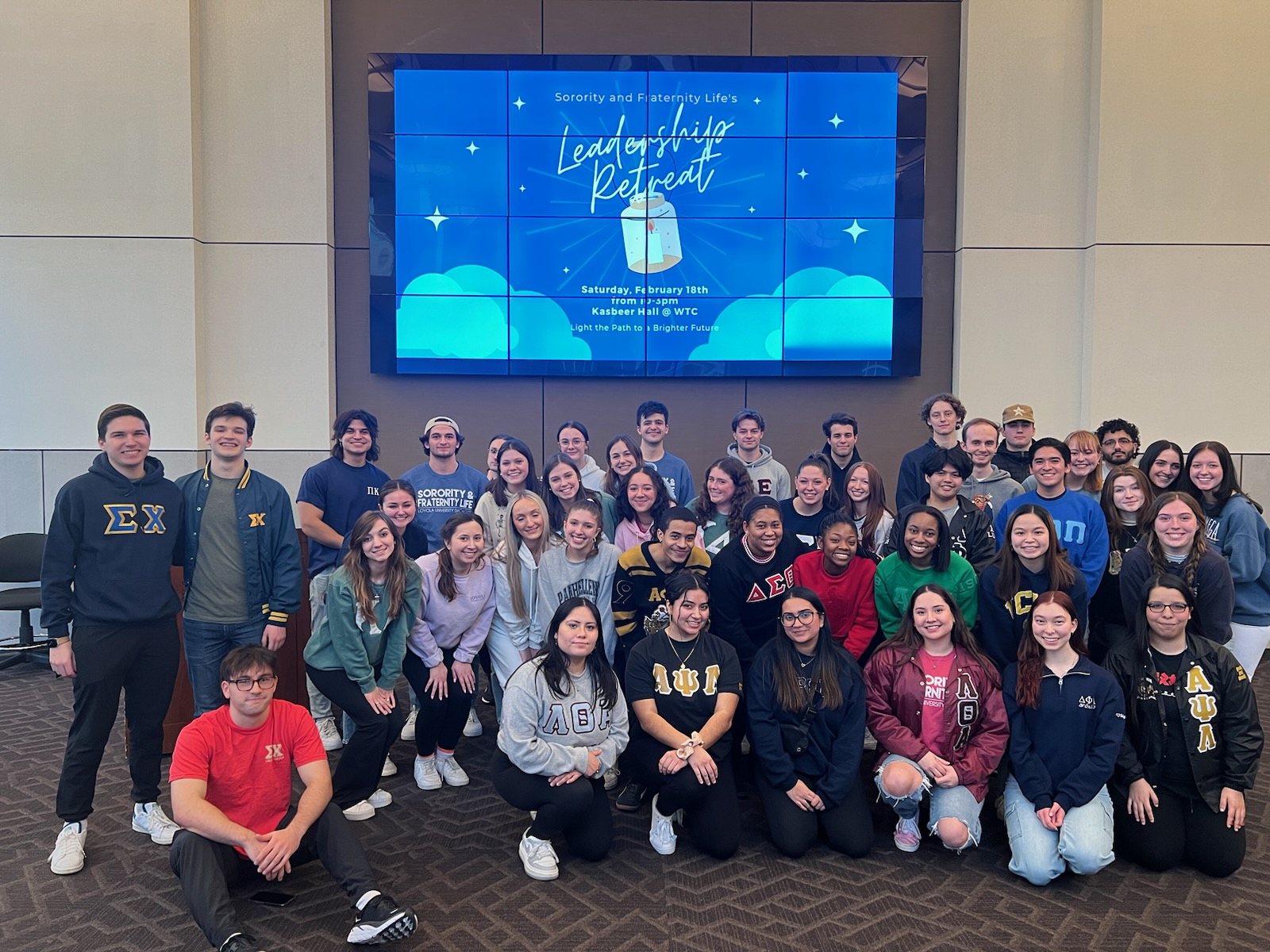 Sorority & Fraternity Life students in a room with tan walls and gray floor standing together in front of a tv screen and kneeling on the floor to pose for a picture